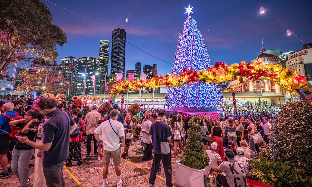 A crowd of people are gathered at a Christmas festival at Fed Square. There is a large glowing Christmas tree.