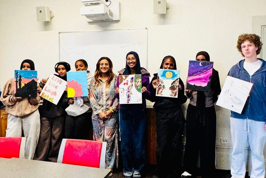 A row of smiling teenagers standing inside a studio space, all holding up artworks they've created. 