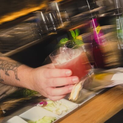 A blurred tattooed arm holding out a cocktail over a bar