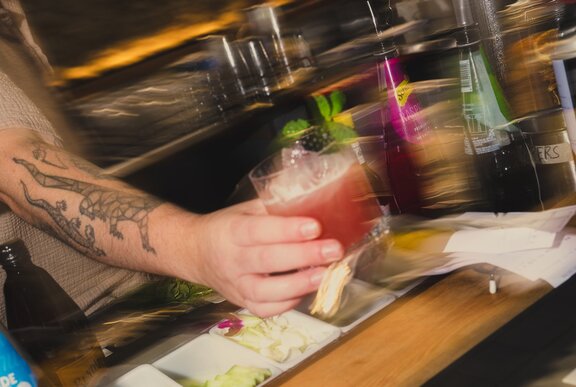 A blurred tattooed arm holding out a cocktail over a bar