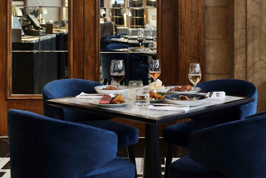 Table set for four, with plates of food and glasses of wine, navy velvet club chairs, inside a wood-panelled dining room.