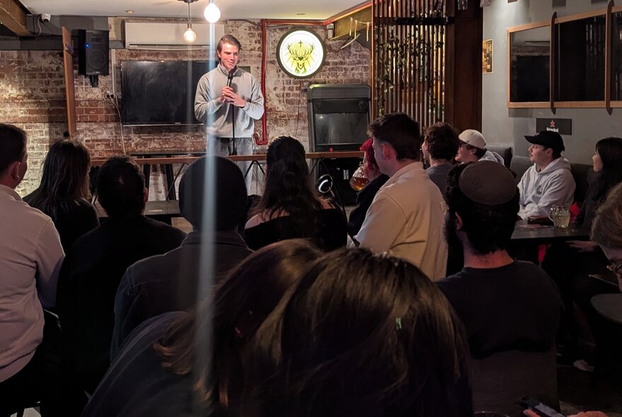 Seated audience watching a standup comedian perform with microphone in a small underground venue.