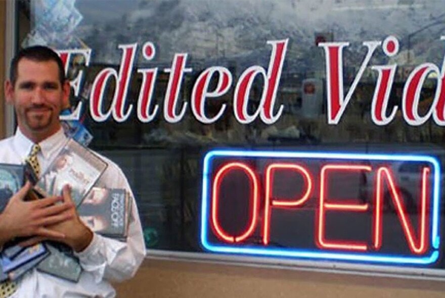 A still form the movie Cleanflix, of a man in a shirt and tie holding many VHS tapes and DVDs in his arms, with a neon shop sign in the background.