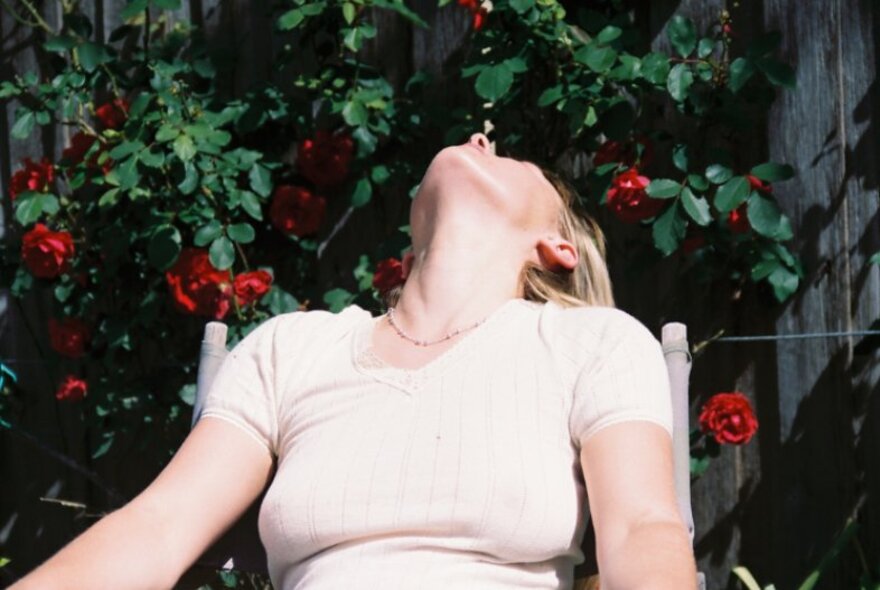 Person wearing a t-shirt leaning very far back in a chair with their head tilted backwards, in a sunny garden. 