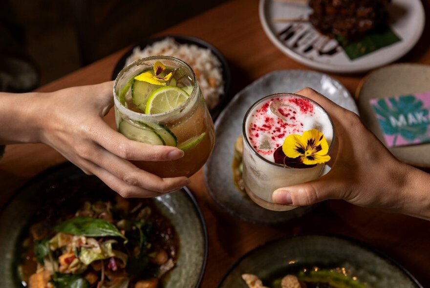 Two hands holding colorful beverages and clinking them together in a celebratory manner above plates of food.