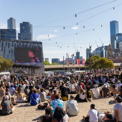 People gathered seated in front of the big screen in Fed Square with sunny blue sky.