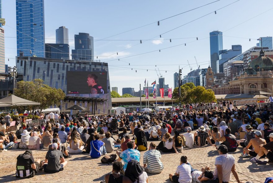 People gathered seated in front of the big screen in Fed Square with sunny blue sky.