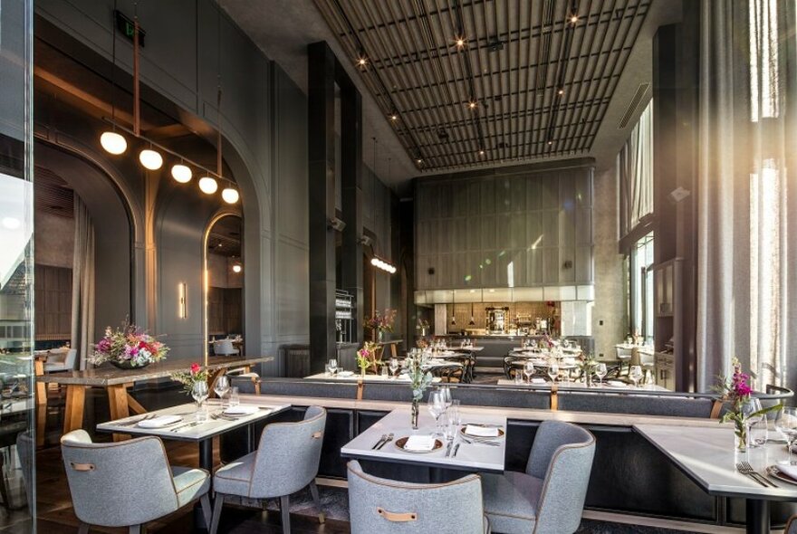 Long view of the interior of a high ceilinged restaurant space with tables of different sizes, upholstered dining chairs, decorative lighting and a panelled ceiling.