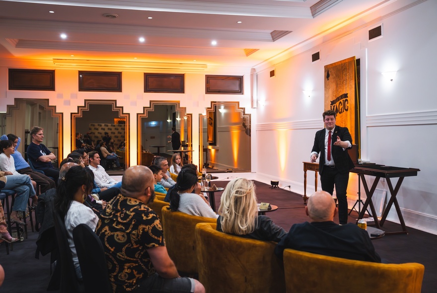 A seated audience in an intimate theatre room setting watching a magician perform.