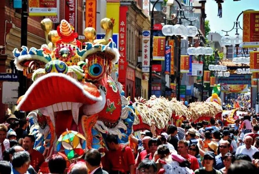 A dragon parade winding through crowd in Chinatown past restaurants and shops.