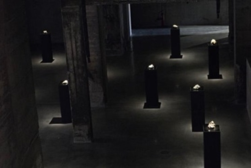 A dark room with 6 narrow display plinths arranged in the space, each with a small object on the top of each one.