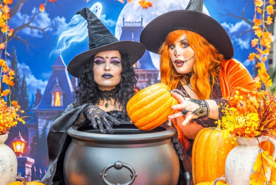 Two women in witch costumes and pointy hats pose with a black cauldron and pumpkins against a spooky castle backdrop.