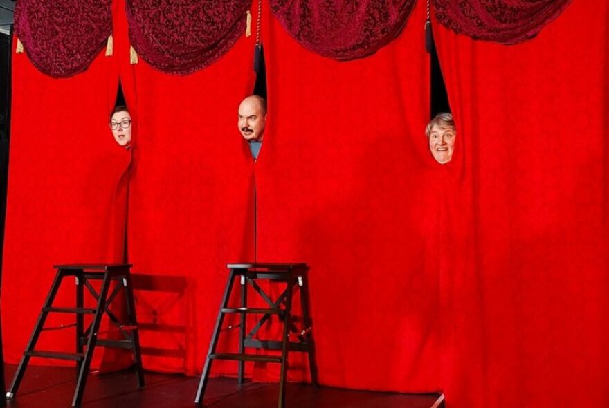 Three faces poking through gaps in a long red curtain on a stage with stools in front of the stage.