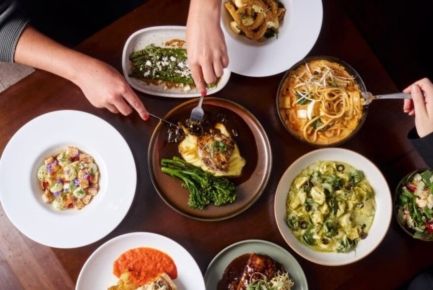 Overhead view of hands with forks selecting food from dishes arranged on a wooden table top.