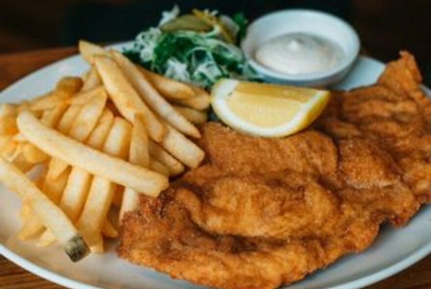 A plate of schnitzel with chips and salad, a slice of lemon as garnish and a small bowl of a condiment on the side.