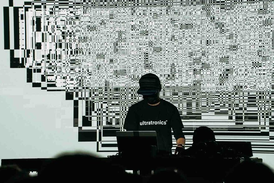 A person in a black t-shirt and cap, behind a laptop, making audio and visuals at the same time, with a screen projecting images in the background.