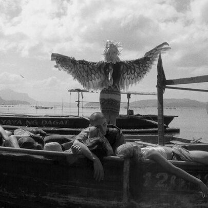Black and white still from the film Essential Truths from the Lake, shows a person standing with their back to the camera on a small boat wearing large, feathered wings with their arms outstretched, with several other people resting or lying down on the same boat.