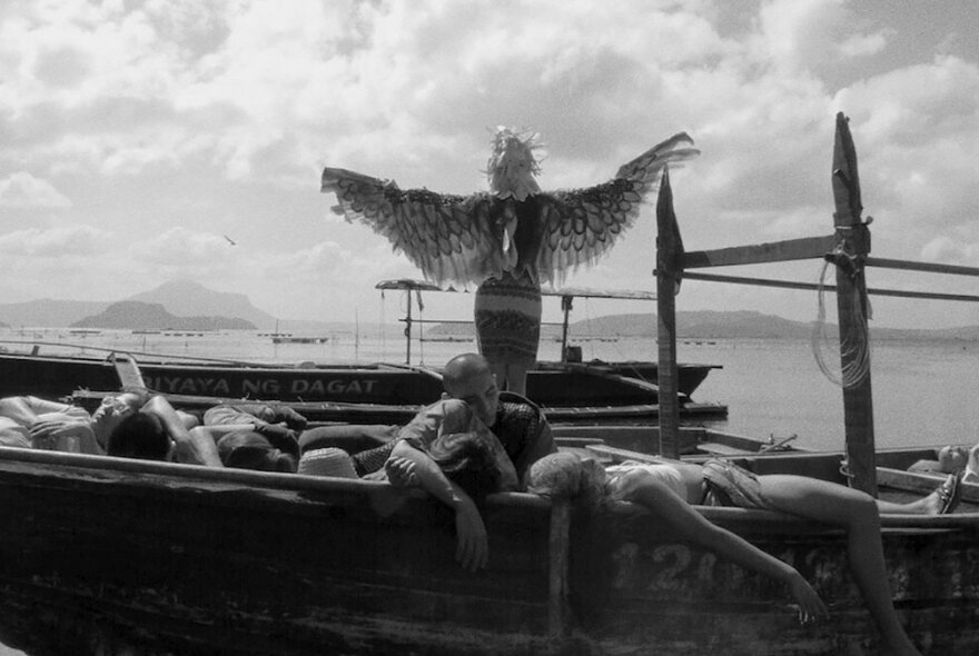 Black and white still from the film Essential Truths from the Lake, shows a person standing with their back to the camera on a small boat wearing large, feathered wings with their arms outstretched, with several other people resting or lying down on the same boat.