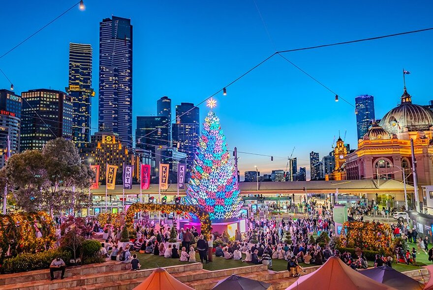 Christmas tree lit up in Fed Square with Flinders Street Station in the background. 