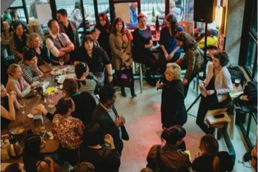 A room full of people casually arranged listening to a central speaker with a microphone. 