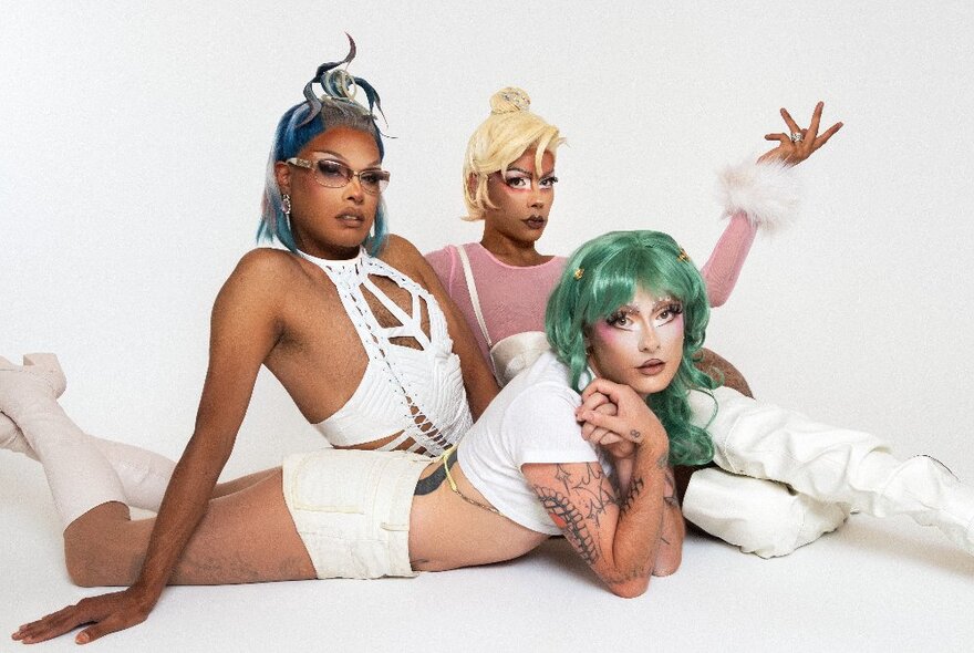 Three drag queens dressed in white outfits and coloured wigs arranged on the floor in glamorous poses. 