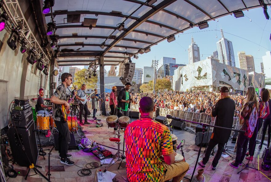 View from the rear of the Fed Square stage of a band playing to a large crowd of people in the piazza, with Fed Square's buildings in the background.