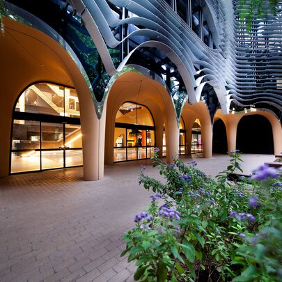 A modern building featuring a ground-floor arcade with large, smooth, light-colored concrete arches and glass entryways, contrasted by a dark, wavy, organic-shaped facade on the upper levels, with green plants and purple flowers in the foreground.