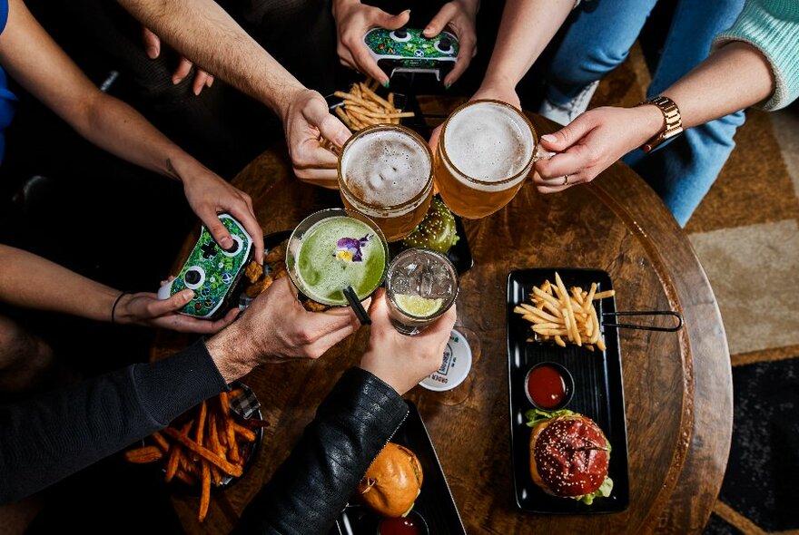 Overhead view of people's hands enjoying drinks and snacks at a low table, some holding PlayStation controllers.