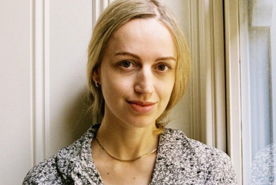 Portrait of a young woman with tied-back blond hair, subtly smiling at the camera with a white panelled background.