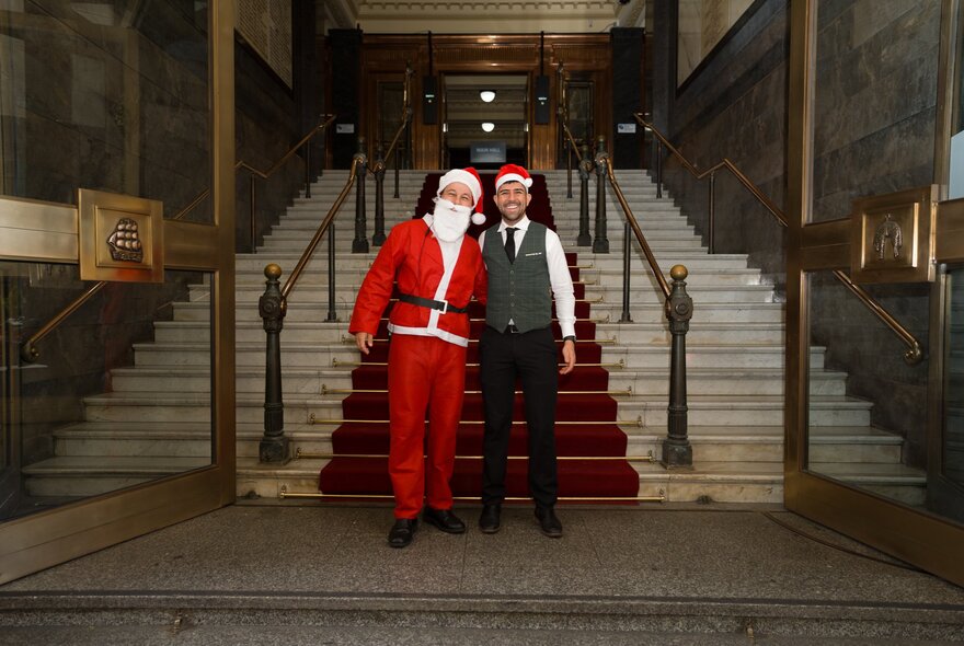 A waiter wearing a Santa hat standing next to a man in a Santa suit at the entrance steps to Melbourne's Town Hall.