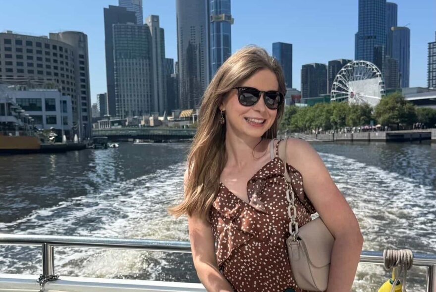 A smiling woman wearing sunglasses, standing on a cruise boat, with the Yarra River and Melbourne city buildings in the background.