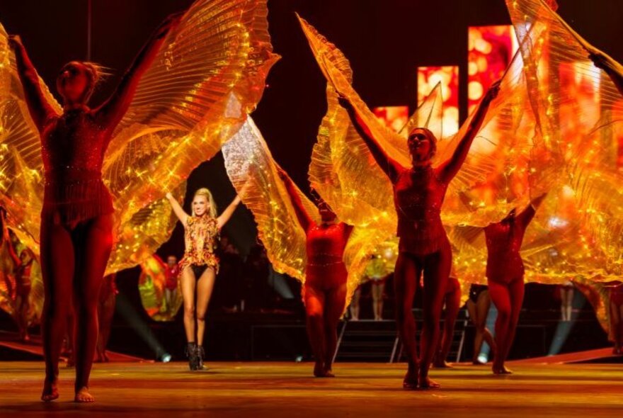 Dance troupe performing with orange transparent wings on stage.