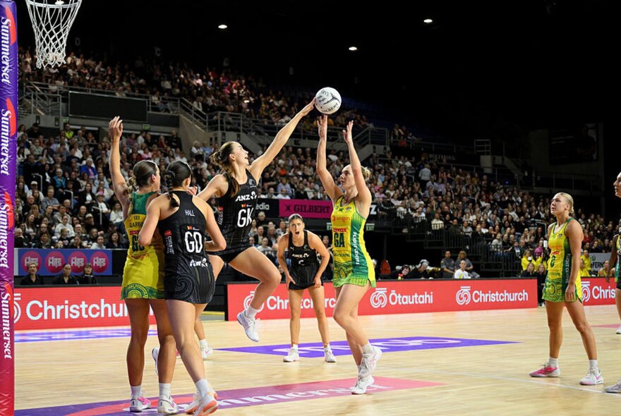 A netball match between the New Zealand Silver Ferns and the Australian Diamonds, with an Australian player throwing the ball towards the netball ring, blurred spectators in the stadium in the background.