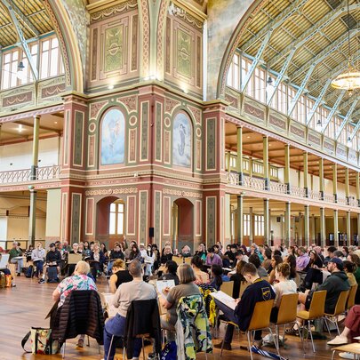A group of people sit on chairs in rows within the ornate, high-ceilinged interior of the Royal Exhibition Building taking part in a drawing workshop.