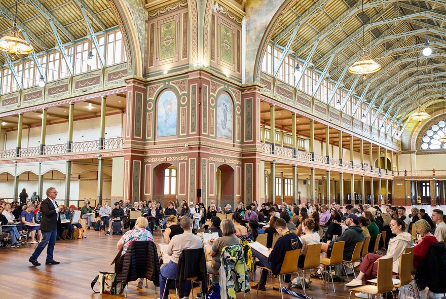A group of people sit on chairs in rows within the ornate, high-ceilinged interior of the Royal Exhibition Building taking part in a drawing workshop.