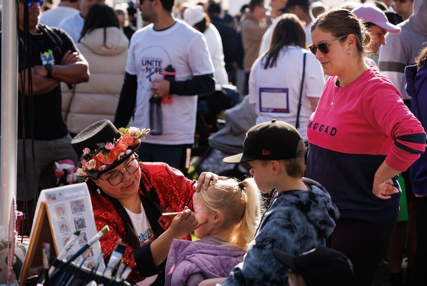 Woman wearing a flowered hat painting a child's face while a woman and boy look on.