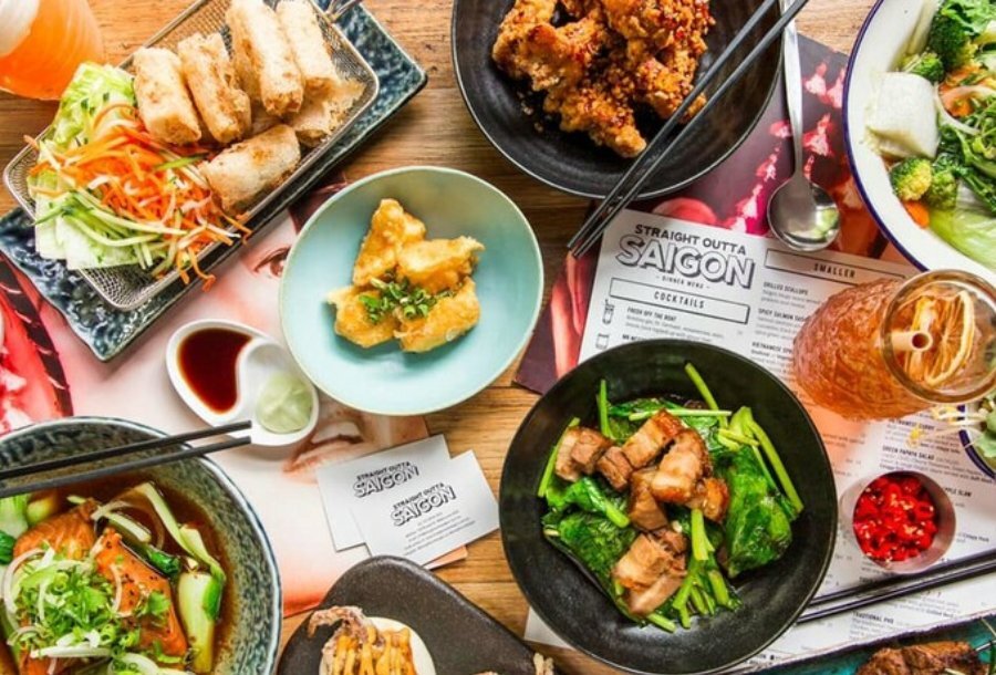 A table with a plate of spring rolls, a plate of fried chicken, a plate of dumplings, a plate of green vegetables and pork belly, a noodle soup and a cocktail. 