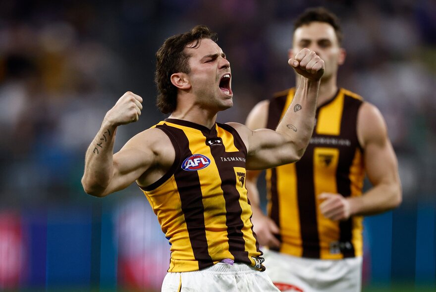 A Hawthorn football player looking jubilant with his arms up in a muscle pose on the football field, another player running behind. 