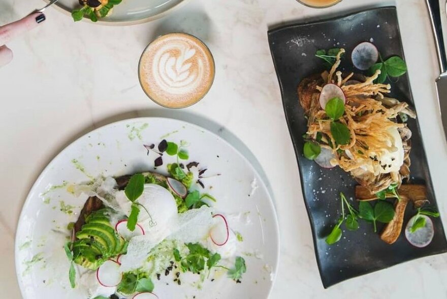 Overhead view of two plates of artfully arranged food and a cafe latte in a glass.