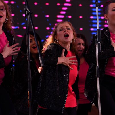 Several performers wearing dark jackets and vibrant pink shirts singing on a stage with microphones as part of a choir.