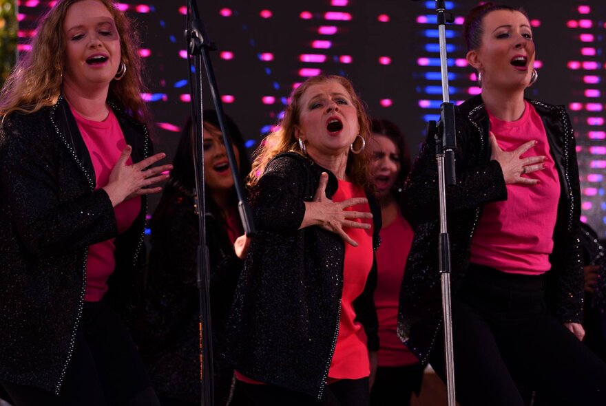 Several performers wearing dark jackets and vibrant pink shirts singing on a stage with microphones as part of a choir.