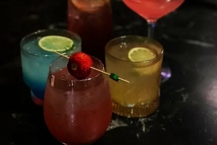 Three cocktails of a varying colour served in short tumblers, one with a cherry garnish.