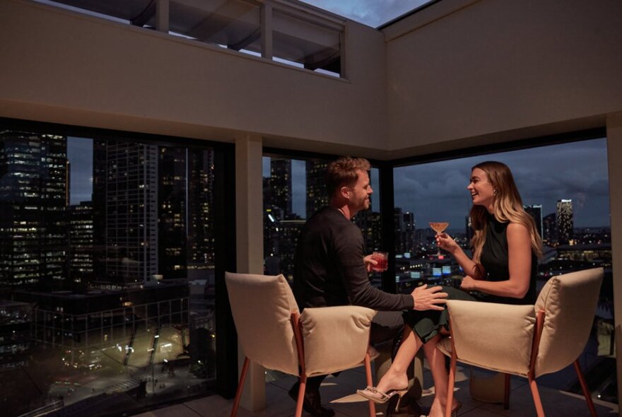 A couple enjoying a romantic drink in a secluded corner of a bar, with windows behind showcasing the city lights at night.