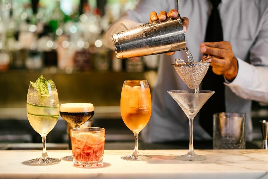 A selection of cocktails on a bar with a bartender pouring a martini.