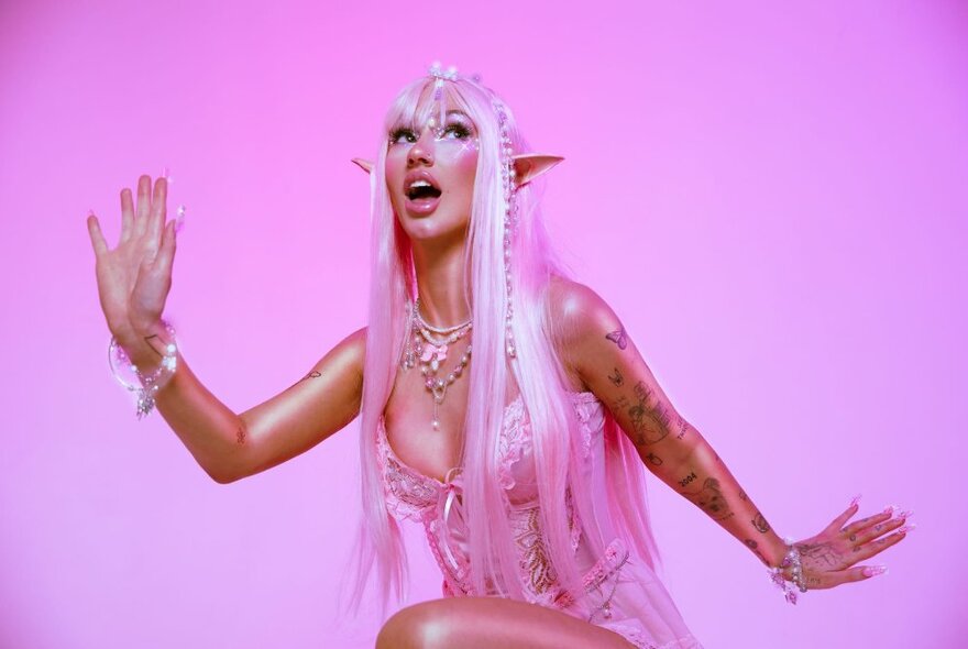 Singer Peach PRC, wearing pixie eras, with long pink hair, jewellery and posing against a pink background.