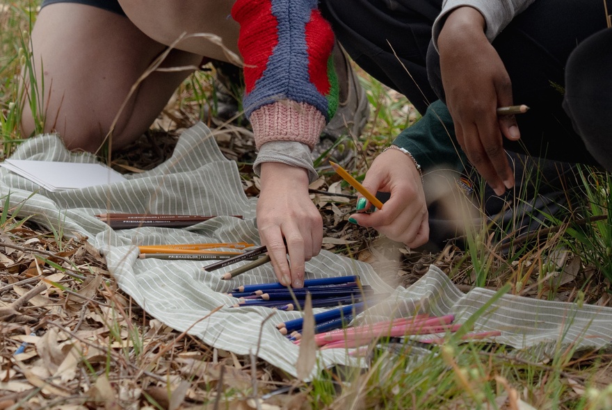 Hands holding coloured pencils on a piece of cloth in an outdoor setting.