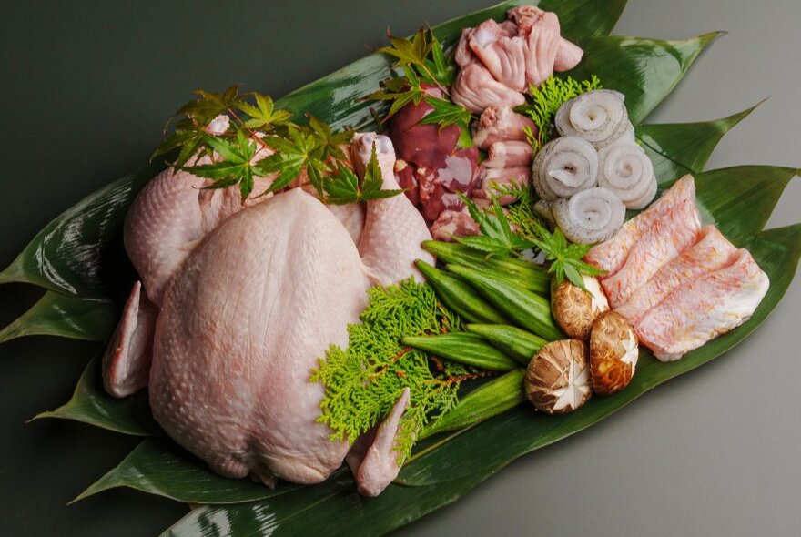 A platter lined in banana leaves with raw meats, including a whole chicken, laid out alongside mushrooms and green leafy vegetables.