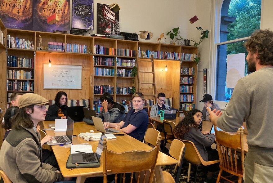 Children seated at tables with laptops, watching a teacher.