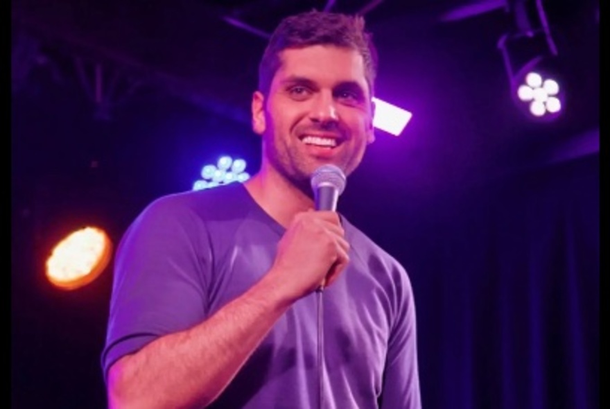 Comedian, Elijah Dries, wearing a purple t-shirt and holding a microphone on stage with stage lighting in the background.