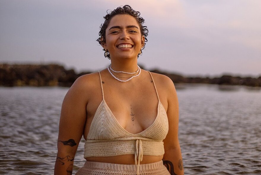 A smiling woman wearing a crochet bikini top in front of a body of water.